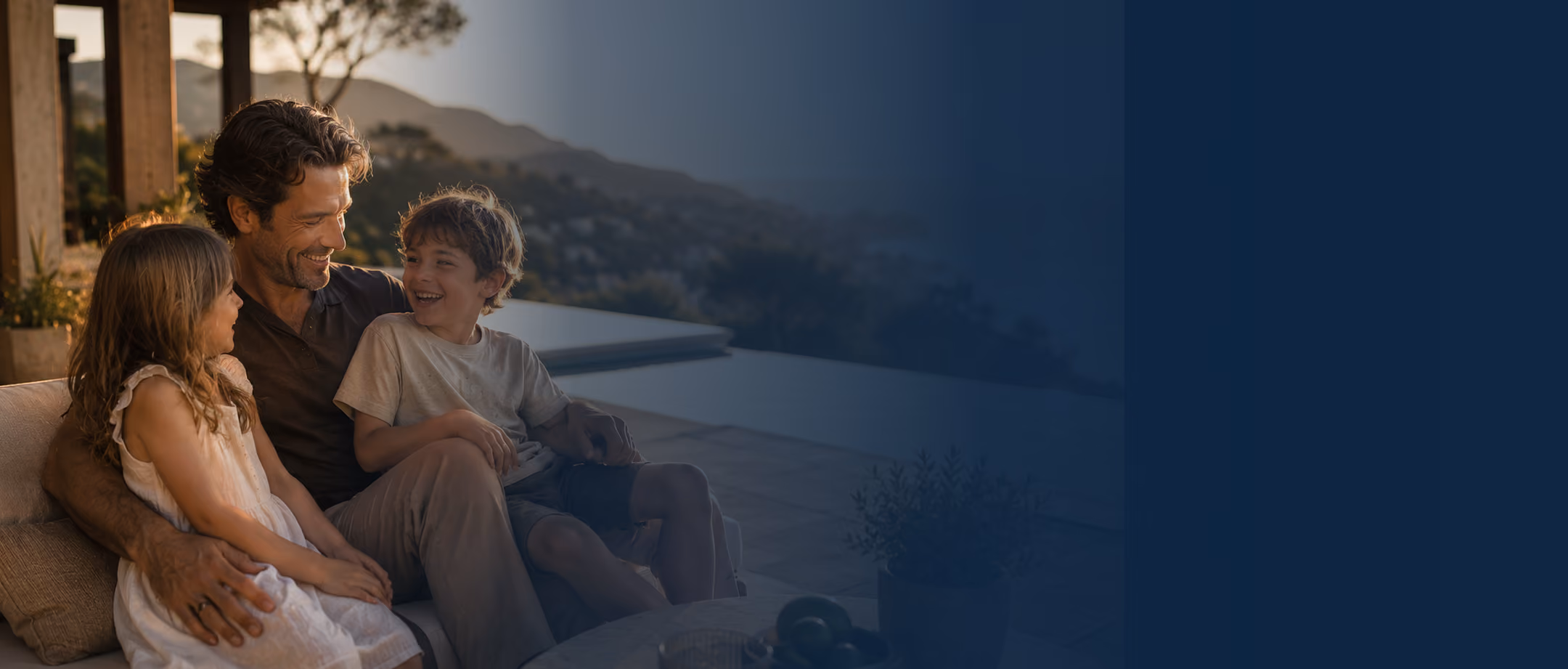 Man sitting outdoors on a sofa smiling with two children on his lap by pool and mountains.