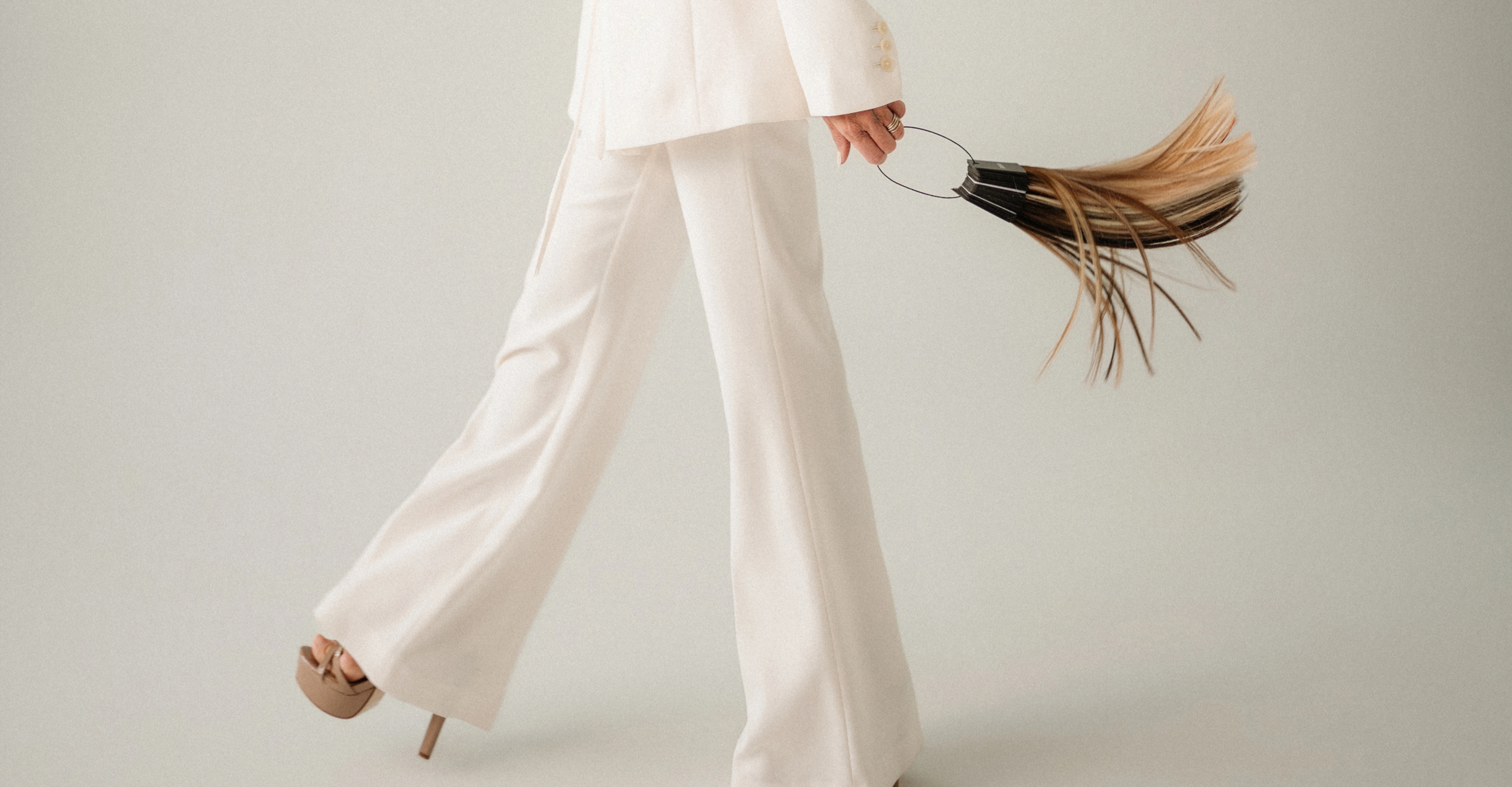 Person in cream pantsuit and heels walking, holding a fan of hair extensions.
