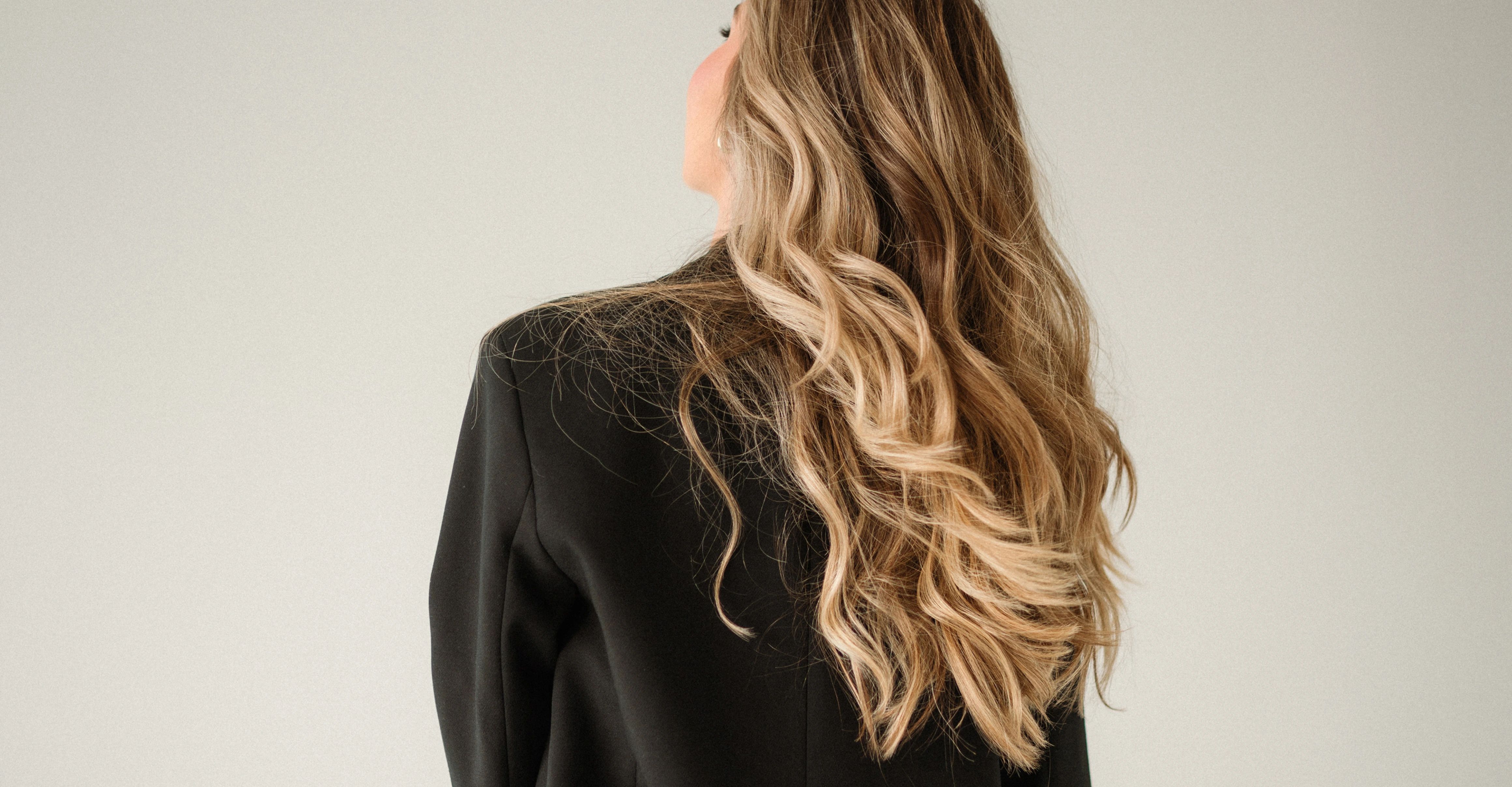 Woman with long, wavy lived-in blonde hair wearing a black blazer turned away from the camera.