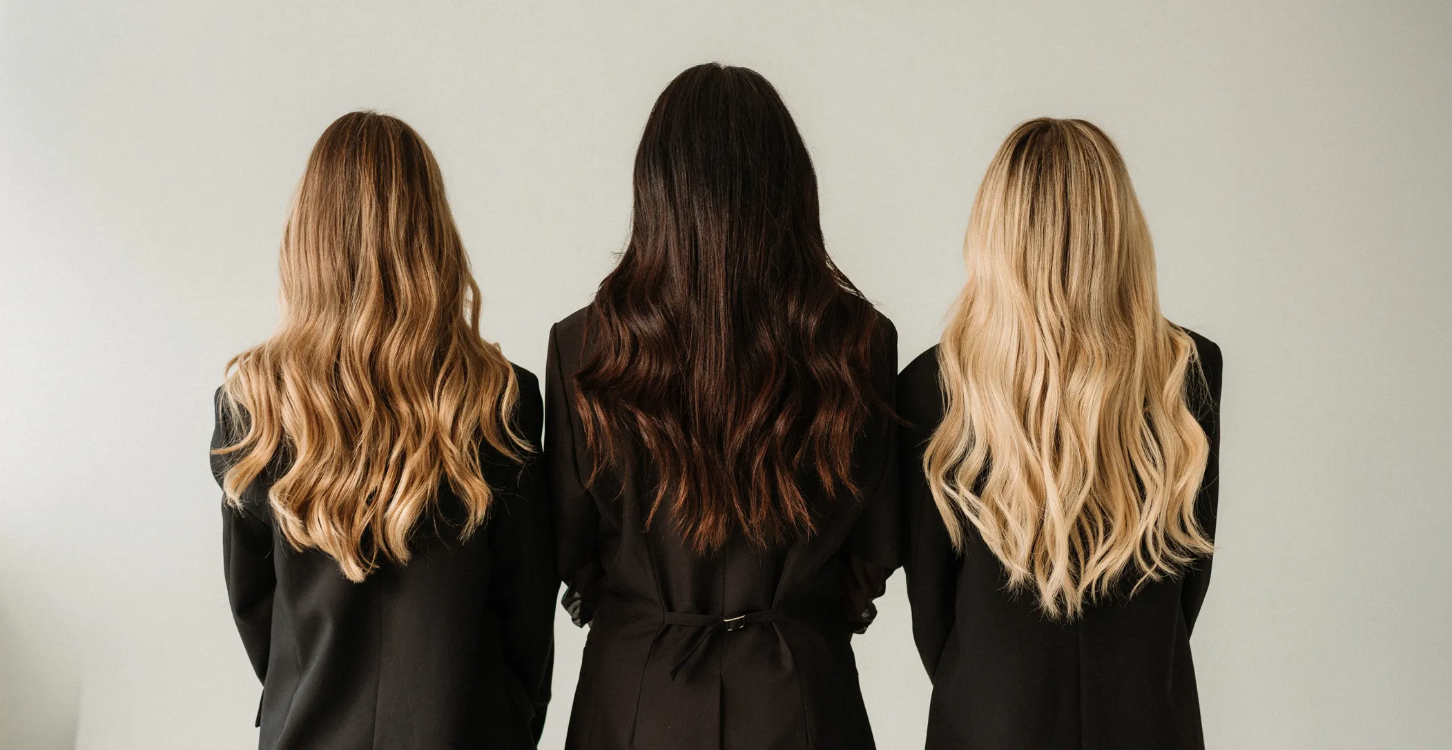Three women with long wavy hair in black outfits stand back to the camera against a plain wall.