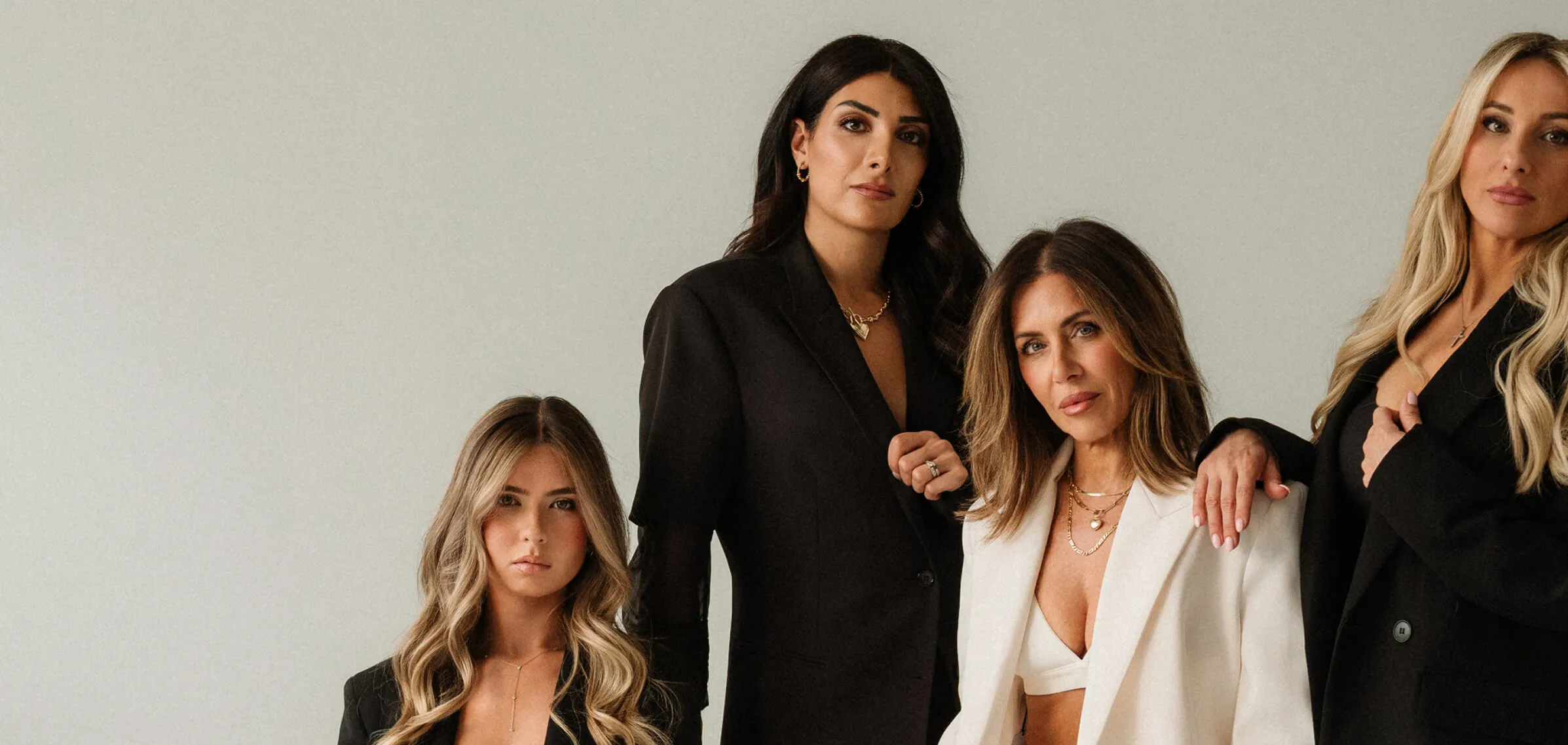 Portrait of four women in black and white blazers posing against a plain light gray background.