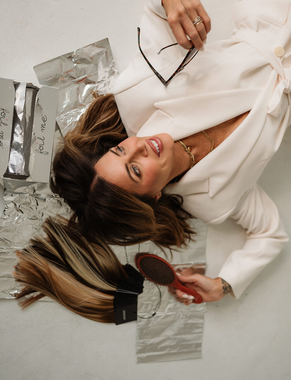 Smiling woman in white blazer lying on floor with hair extensions, foil, hairbrush, and glasses.