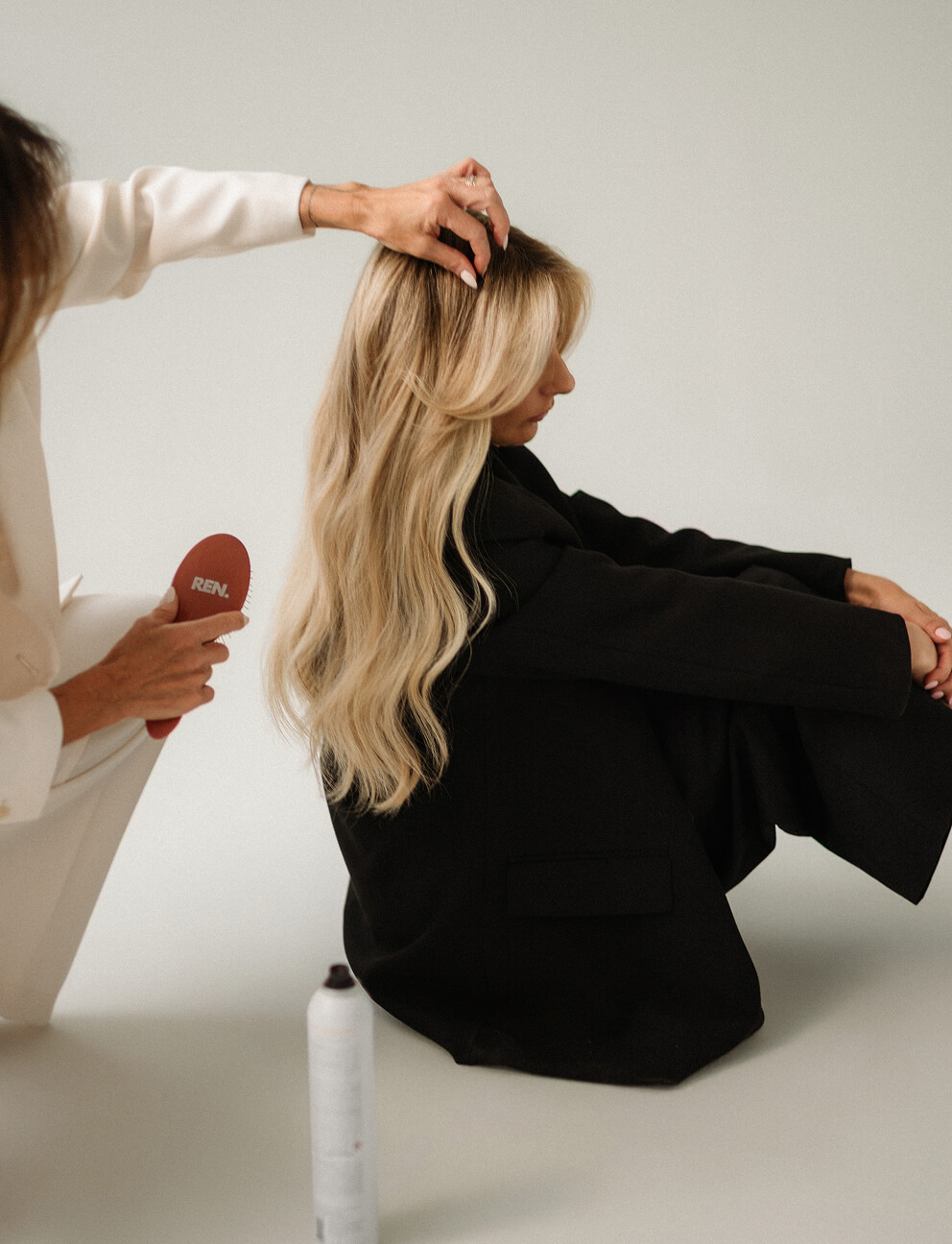 Blonde woman in black jacket seated while another person brushes her hair with a REN brush.