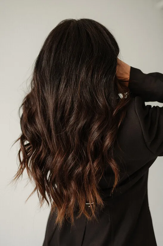 Woman with long, wavy dark brown hair and a black blazer, facing away and holding her head.