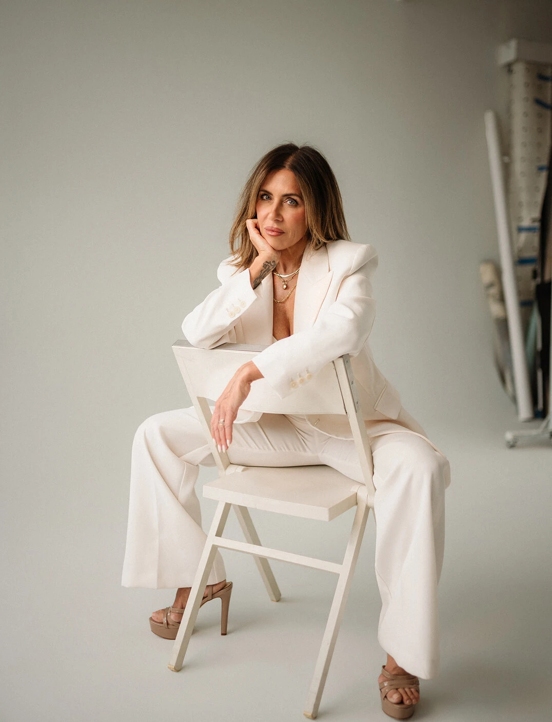 Woman in a white suit sitting backward on a white chair with hand resting on her face.