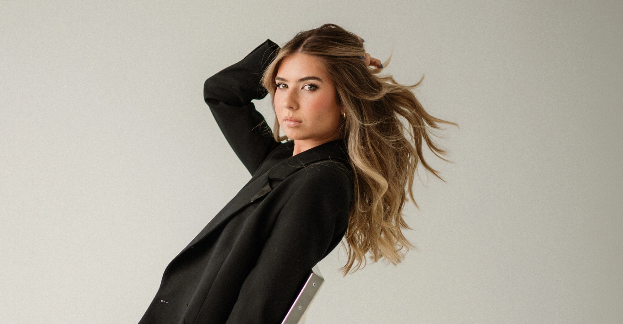 Young woman in black blazer with flowing light brown hair posing against a plain background.