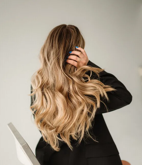 Person with long, wavy blonde hair wearing a black jacket, facing away and touching their head.