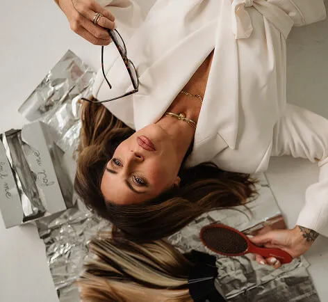 Woman in white blouse with gold necklace holds glasses and brush with hair extensions on shiny surface.