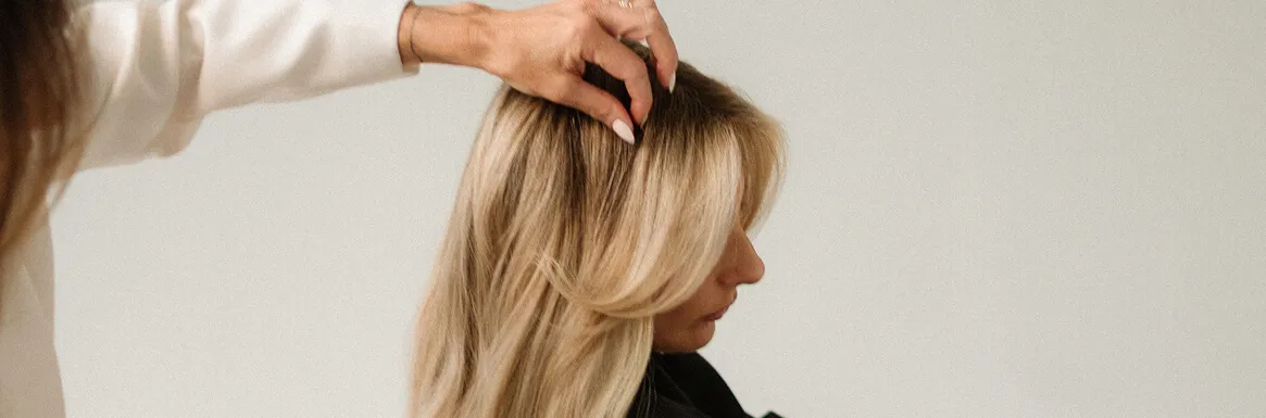 Hand styling long blonde hair of a woman with side part against a plain background.