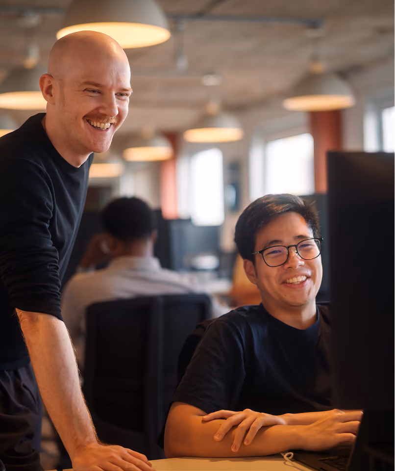 Two men smiling and working together at a computer in a modern office with hanging lights.
