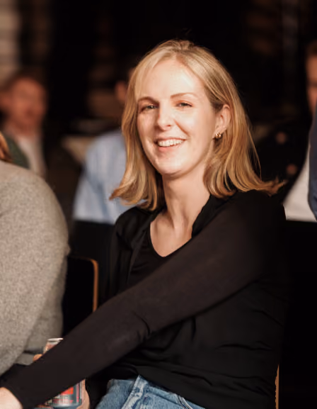 Smiling blond woman wearing black shirt sitting indoors among people in a dimly lit room.