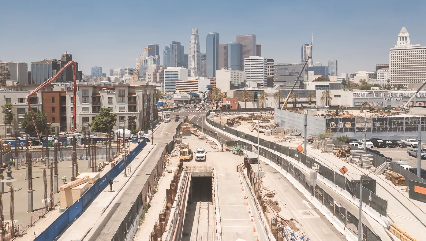 Urban construction site with workers and cranes, city skyline in the background under clear sky.