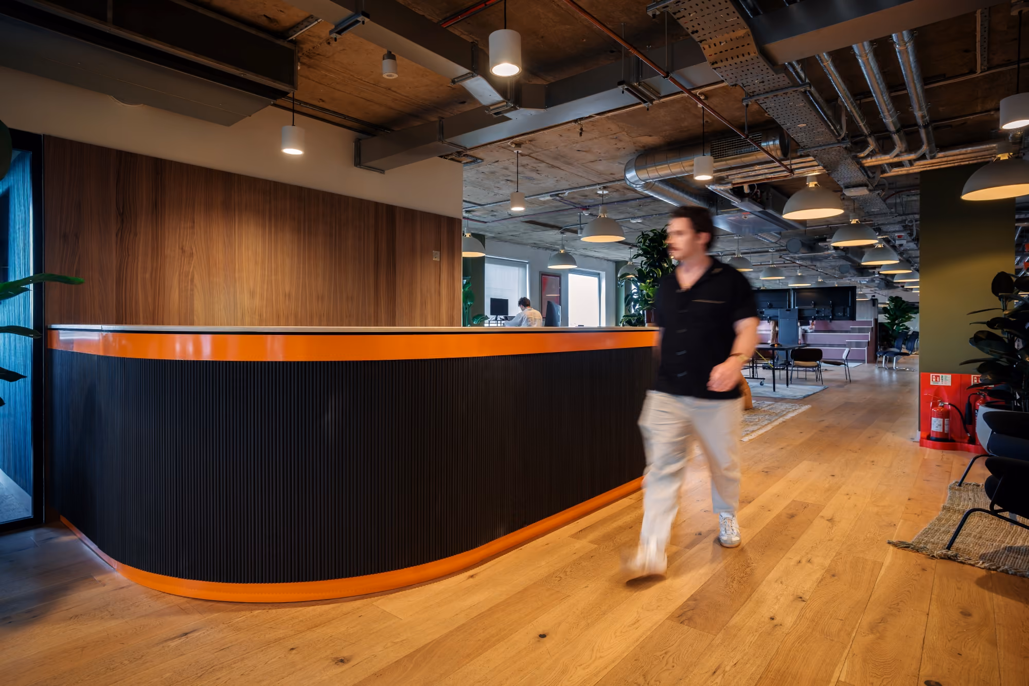 Modern office lobby with orange-accented reception desk and a man walking on wood floor.