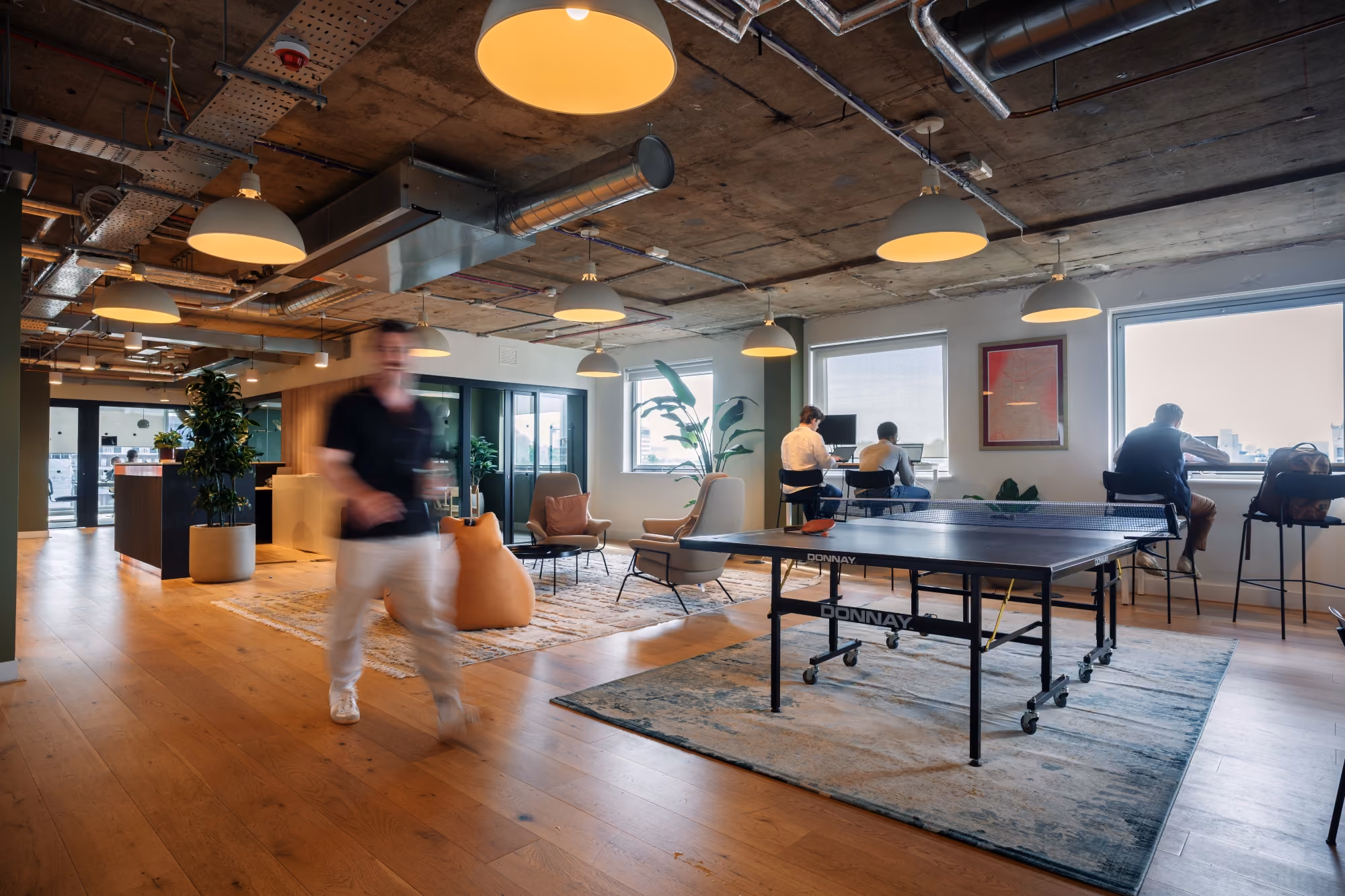 Modern office lounge with ping pong table, seating area, and people working near large windows.