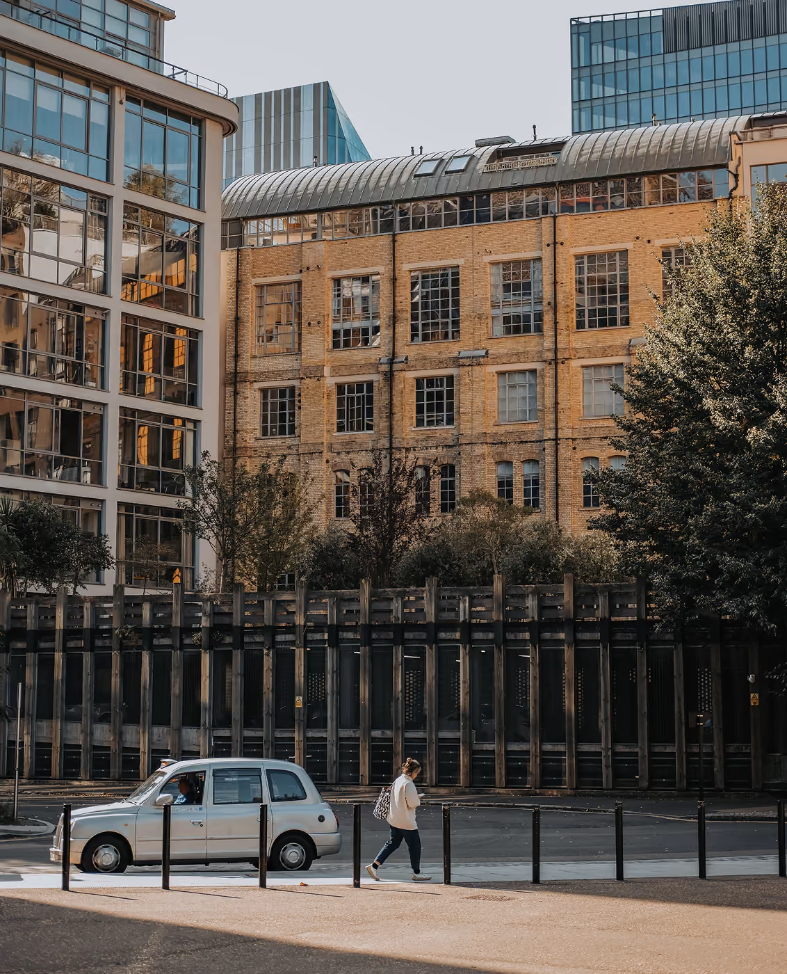 Person walking on sidewalk near a silver taxi with modern and old buildings in the background.