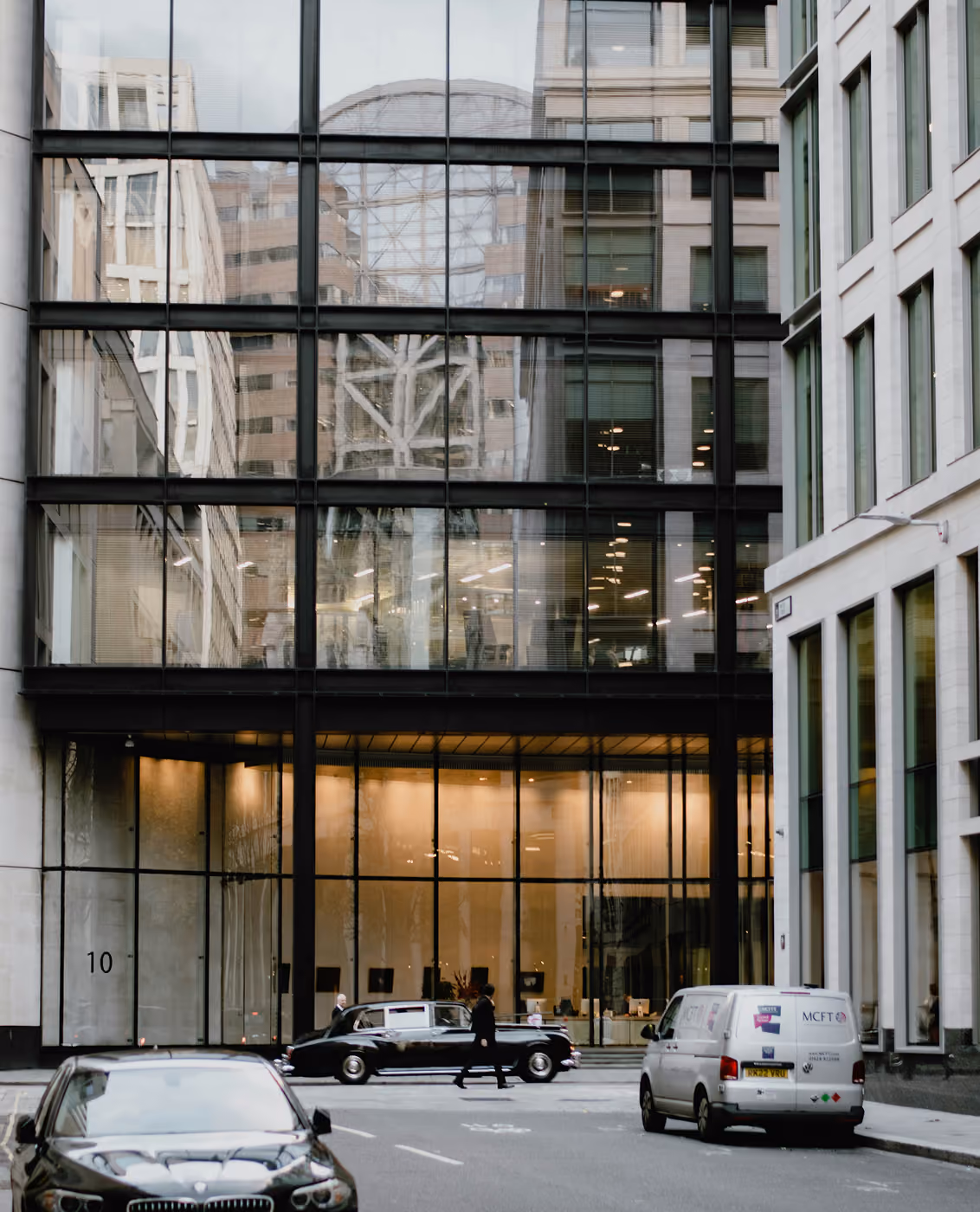 City street view with modern glass building, cars parked and a person crossing the road.