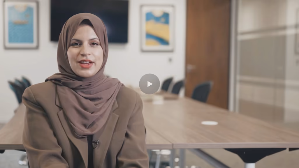 Woman in a brown hijab and blazer speaking in a modern conference room with a wooden table.