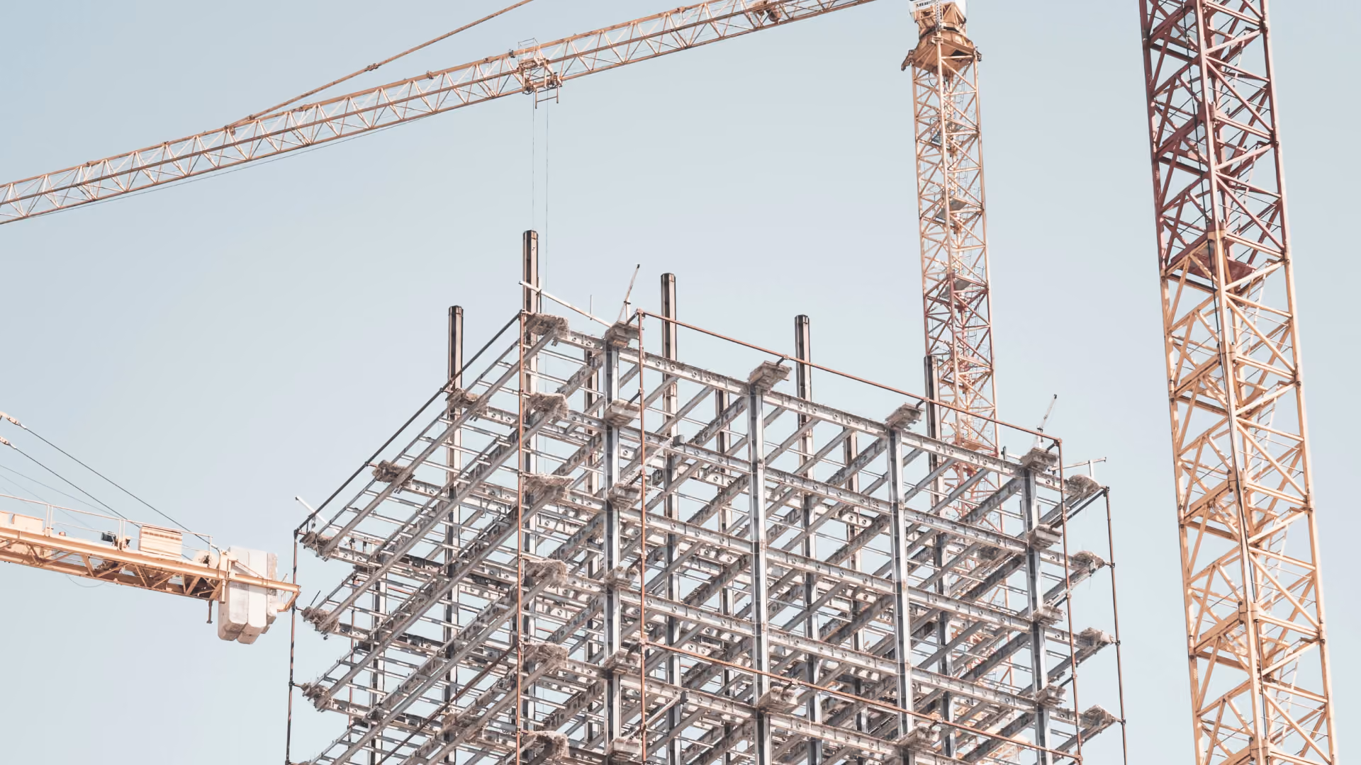 Steel framework structure under construction with two yellow cranes against a clear sky.