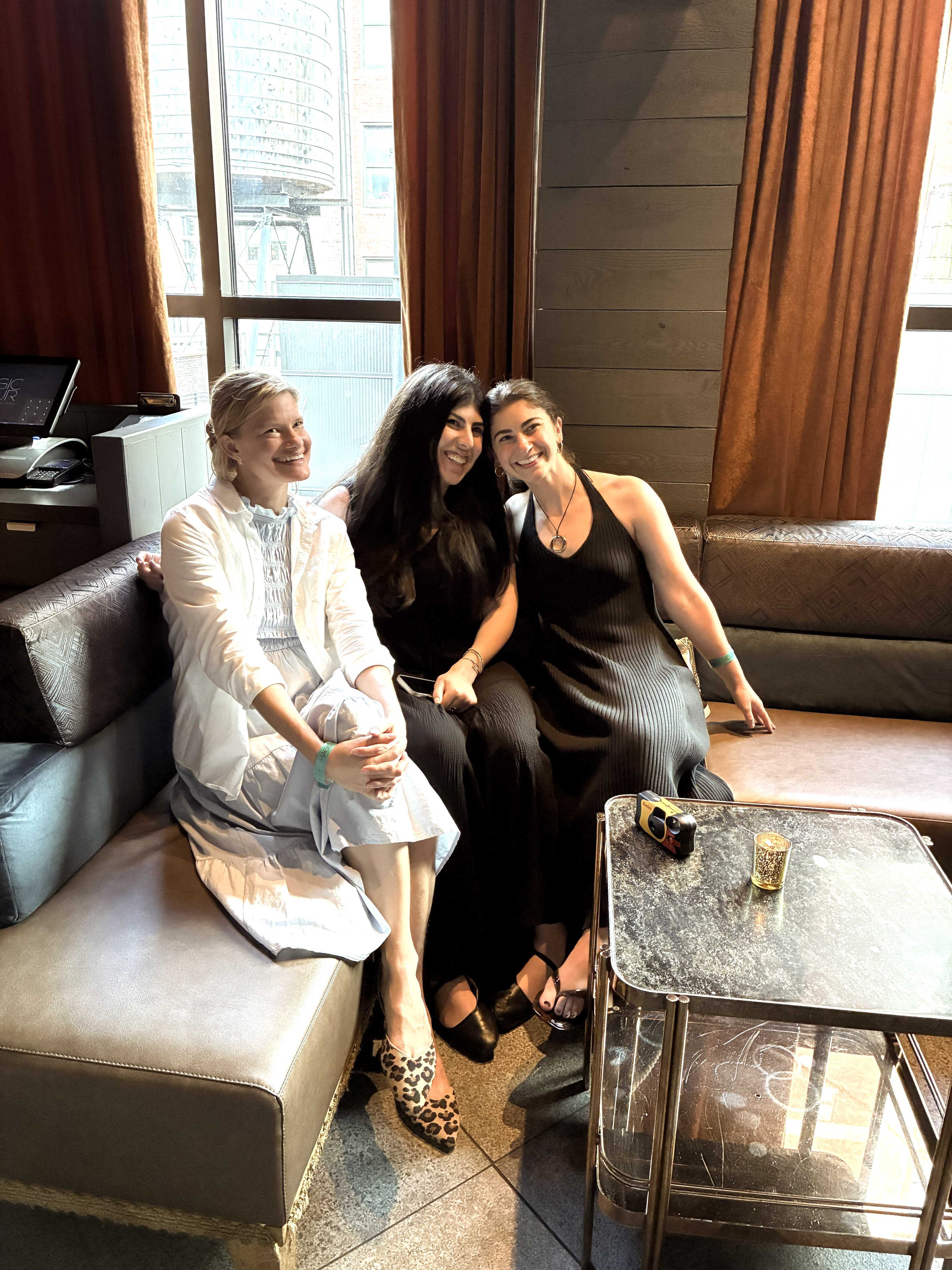 Three women sitting close together on a couch smiling inside a room with large windows and brown curtains.
