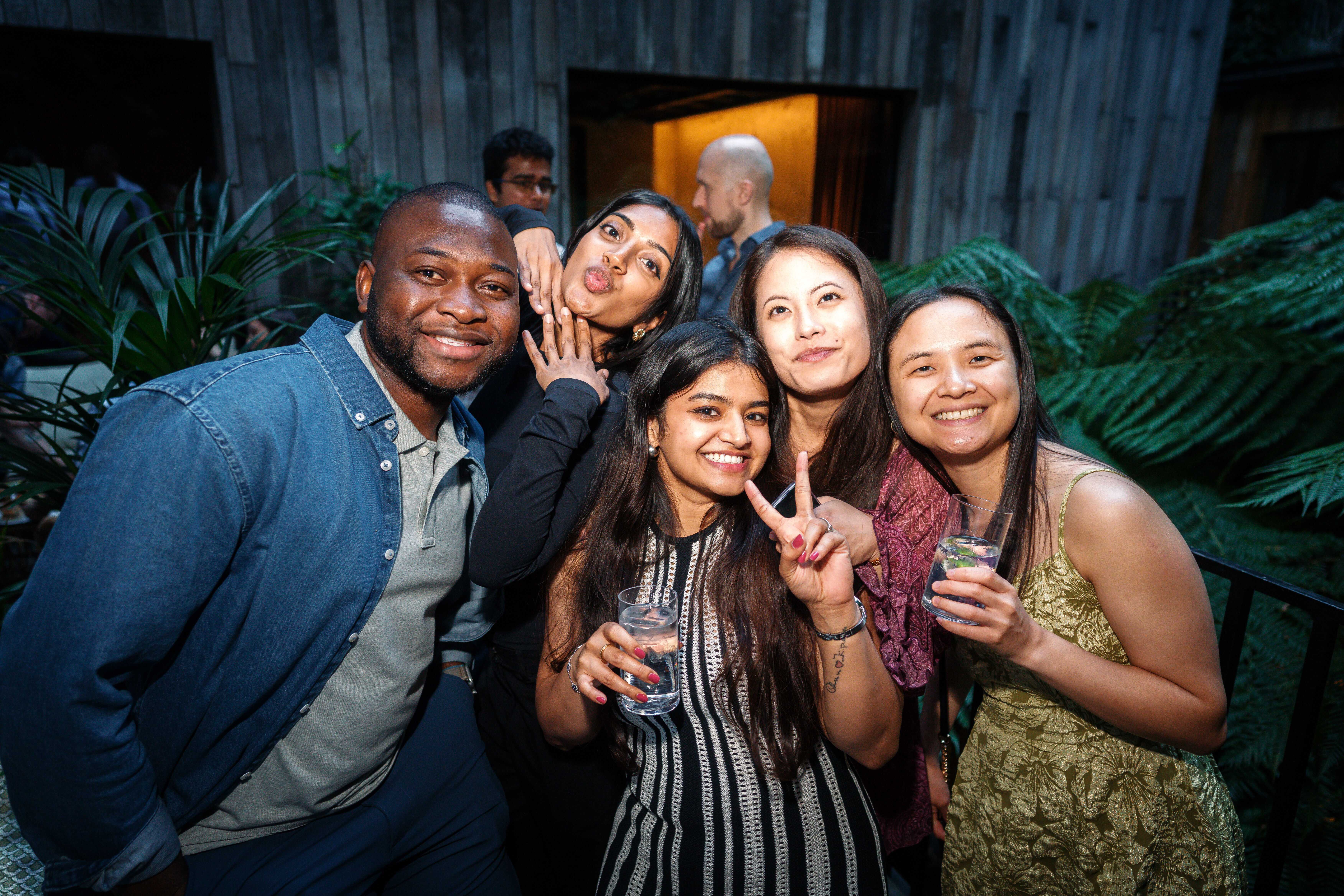 Five people smiling and posing together outdoors at night, two holding drinks and one showing a peace sign.
