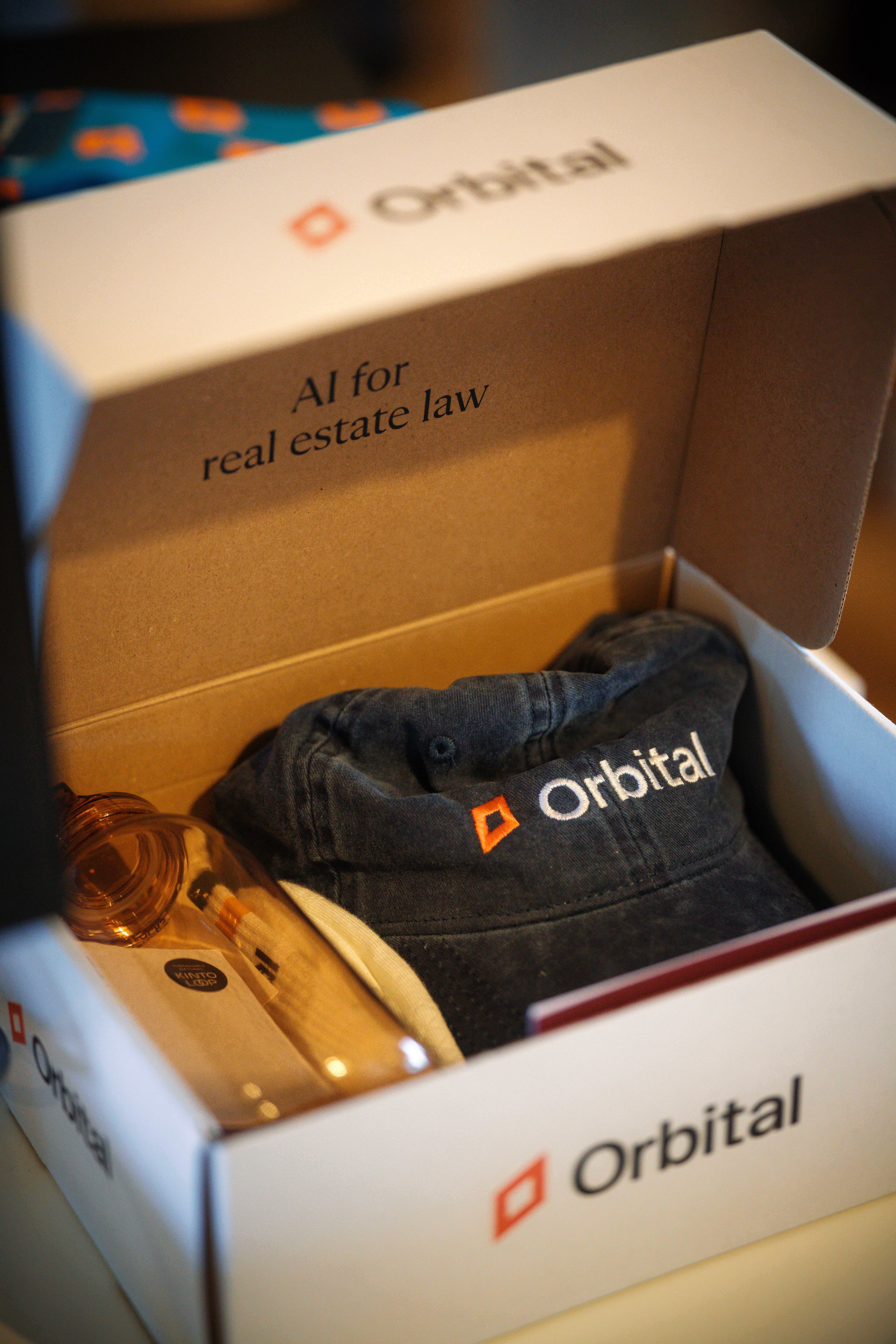 Open box with Orbital branded cap and a transparent water bottle, text inside reads AI for real estate law.