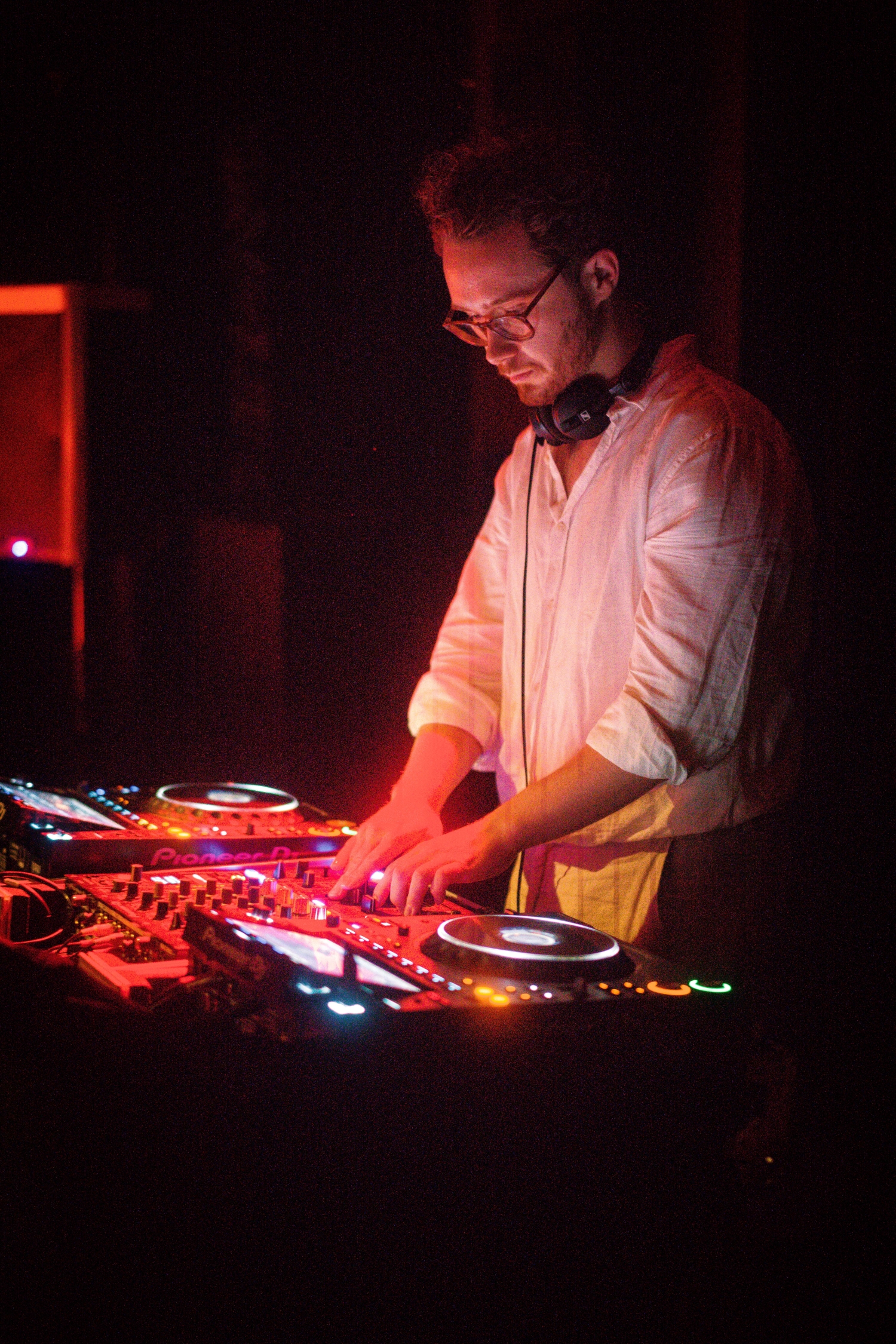 Man DJ wearing glasses and headphones adjusts controls on a Pioneer DJ mixer in dim lighting.