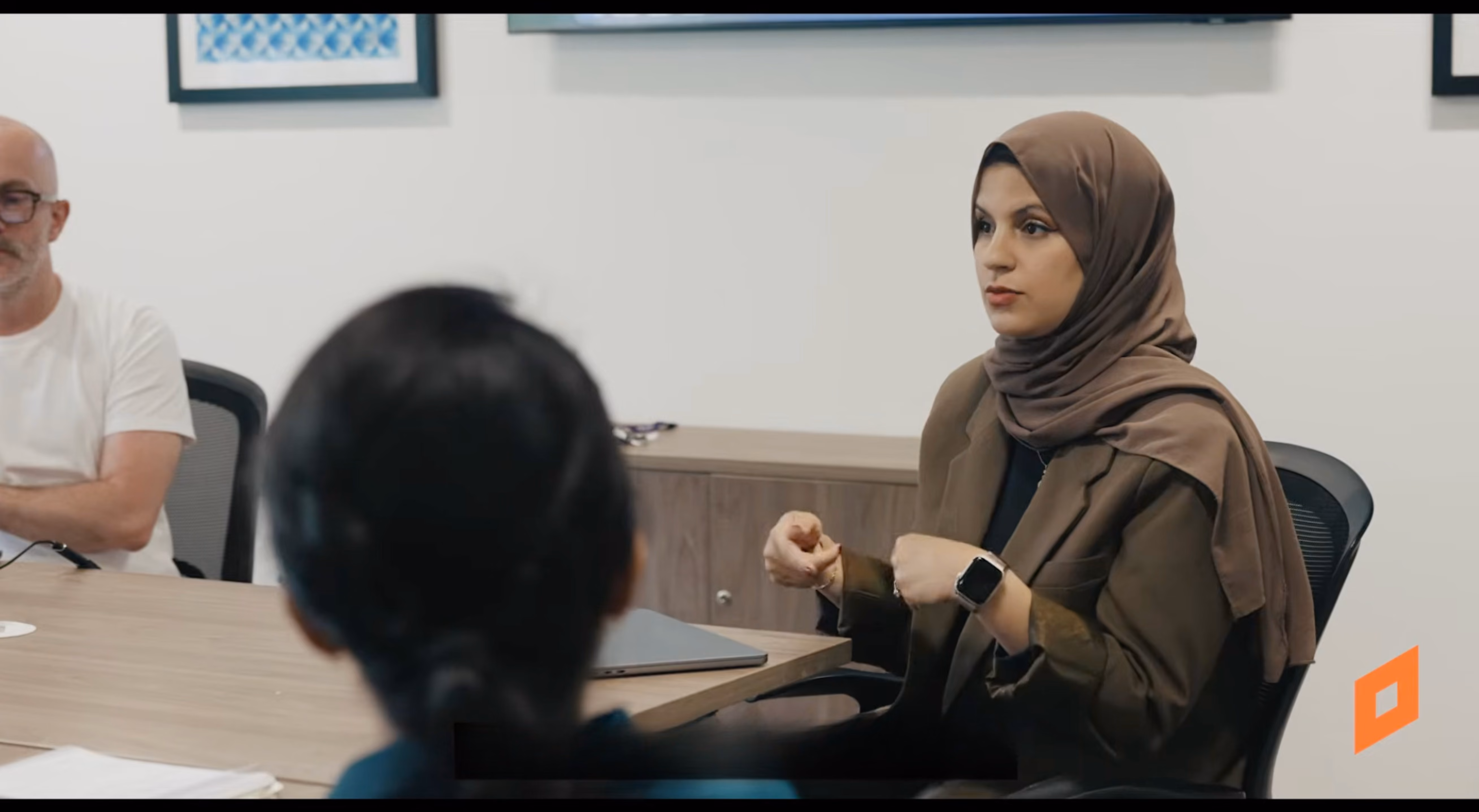 A woman wearing a brown hijab and matching jacket is speaking at a meeting, seated at a conference table with a closed laptop, with two people listening around the table.