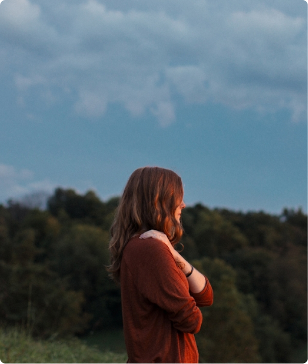 Woman with long hair in a rust sweater standing outdoors at dusk with trees in background.