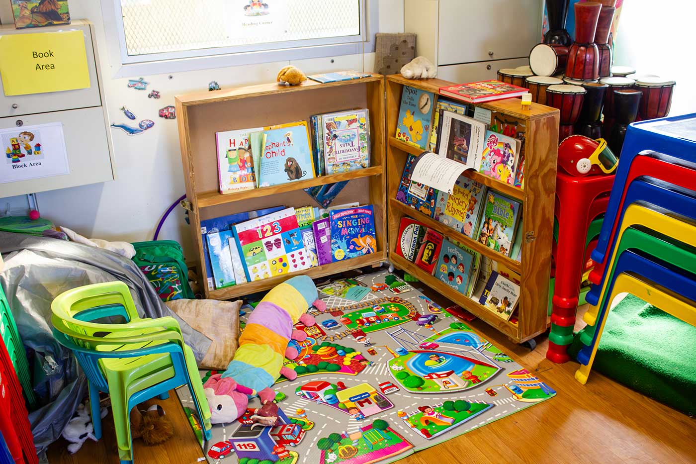 Colorful children's reading area with bookshelves, a play mat, stuffed toy, and stacked chairs.