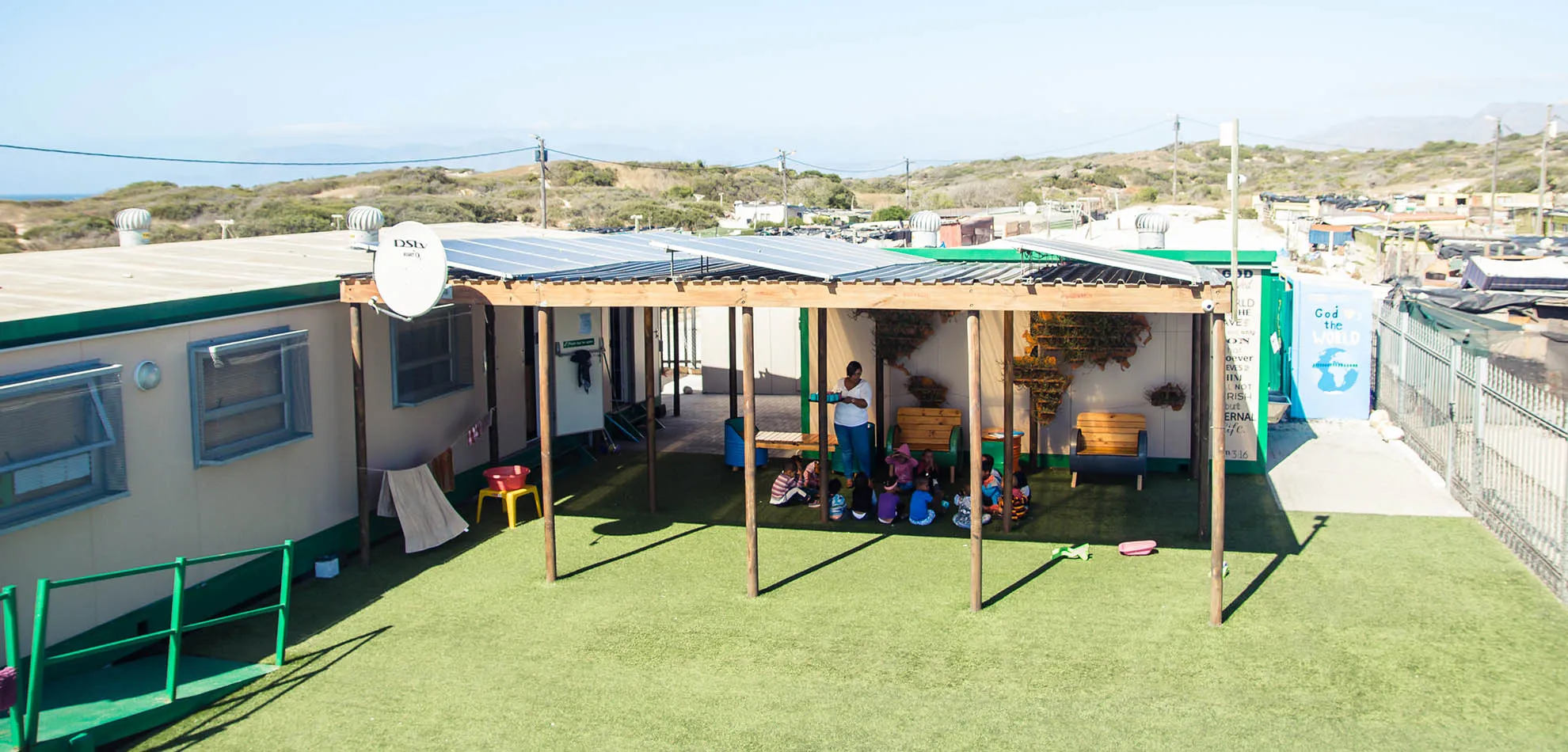 Outdoor classroom with children sitting under solar panel roof and teacher standing nearby.