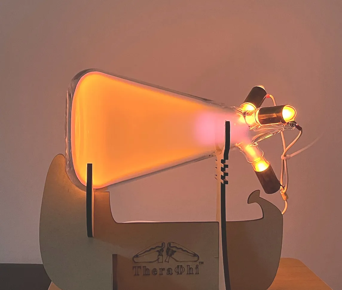 A Theraphi plasma tube glowing warm amber on its wooden stand against a soft pink wall