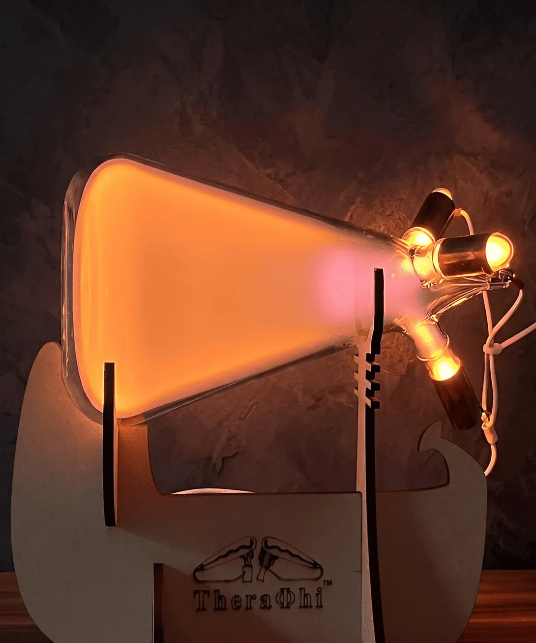 A TheraPhi plasma horn tube glowing warm amber and pink on a wooden stand against a dark textured wall