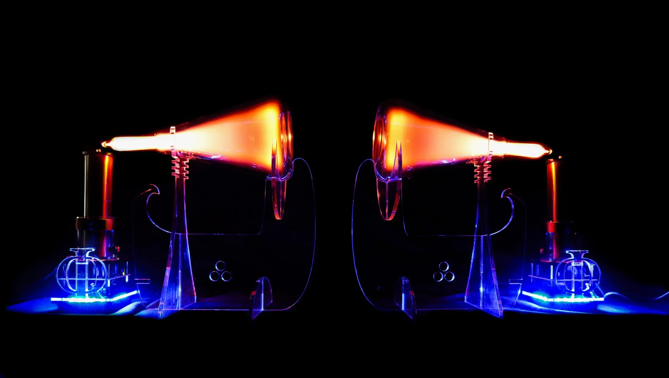 A mirrored pair of glowing plasma flame-tube devices on clear acrylic stands in the dark