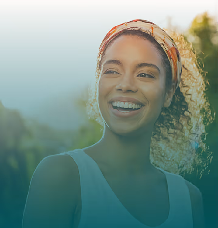 A close-up of a young woman with curly hair wearing a colorful headband, smiling brightly outdoors with sunlight filtering through the trees behind her.