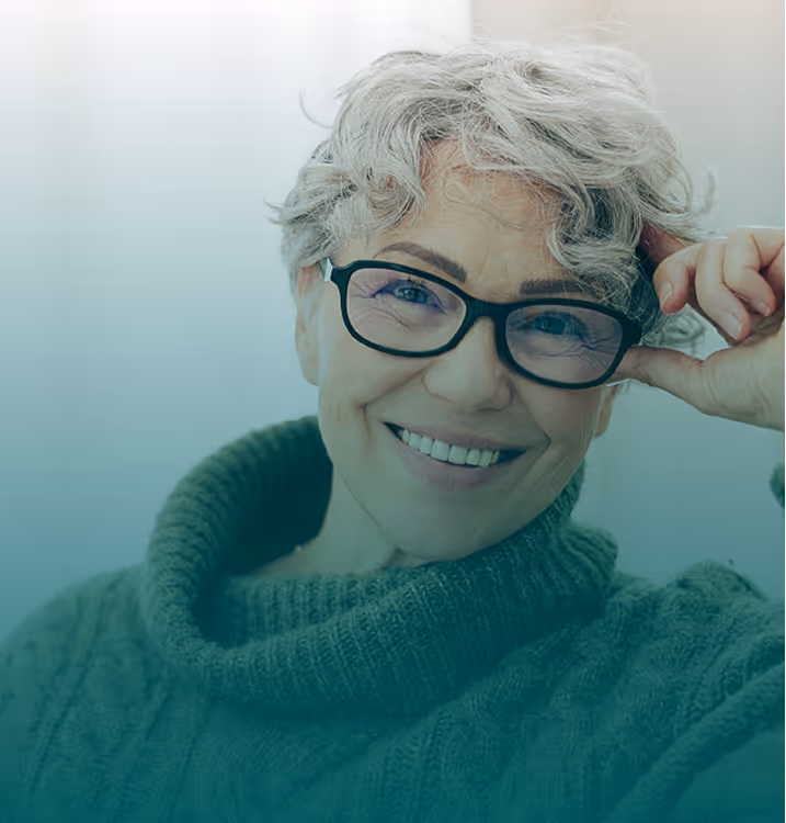 A smiling mature woman with short grey hair and glasses looks directly at the camera while wearing a cozy green cowl-neck sweater, representing healthy and proactive screening practices at every age.