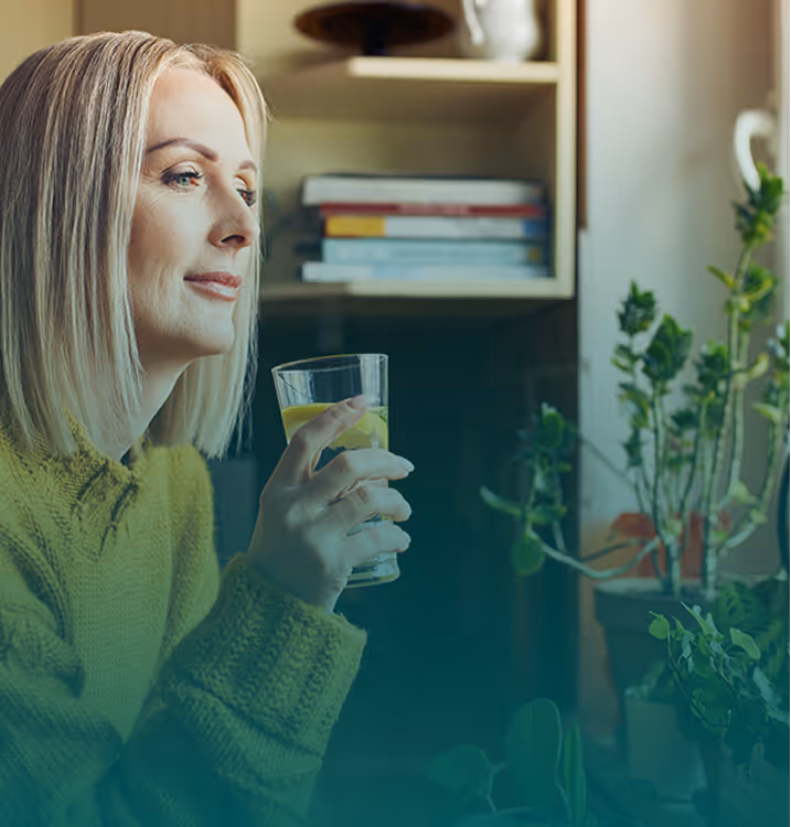 A woman looking out a window while holding a glass of water, surrounded by indoor house plants in a warm kitchen setting.