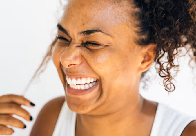 Woman in a white tank top laughs joyfully with eyes closed while playing with her curly hair.