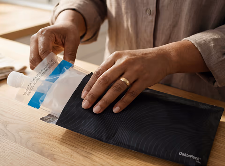 A close-up shot of a person’s hands in a kitchen, carefully placing a sealed, transparent biohazard specimen bag containing a collection vial into a discreet return mailer envelope for an at-home cervical cancer screening test.
