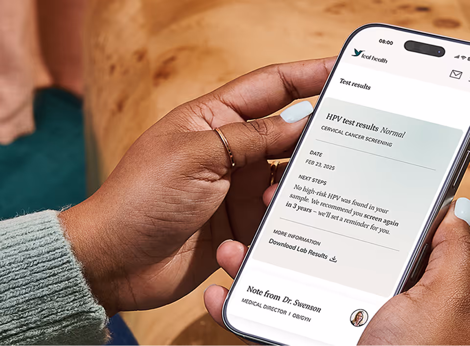 An over-the-shoulder view of a woman sitting on a couch, holding her smartphone to review her secure HPV test results on the screen.