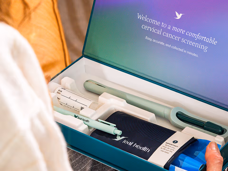 An over-the-shoulder view of a woman unboxing her Teal Health at-home cervical cancer screening kit. The opened box displays the Teal Wand device, a collection vial, a return mailer envelope, and an inside lid reading "Welcome to a more comfortable cervical cancer screening."
