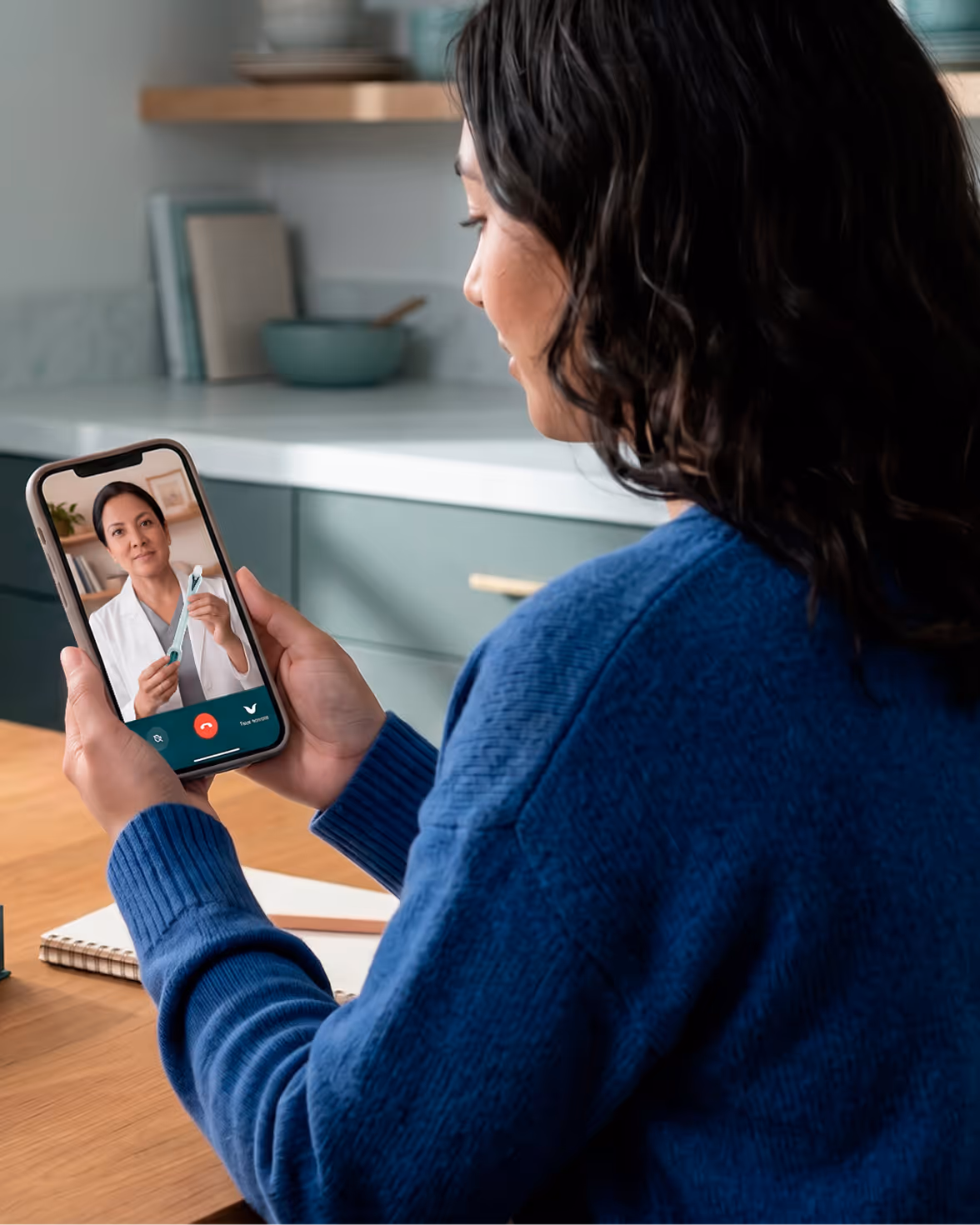 An over-the-shoulder view of a woman sitting at a wooden table, holding a smartphone during a live video telehealth appointment. On her phone screen, a female doctor in a white coat holds a cervical screening device to guide her through the process.