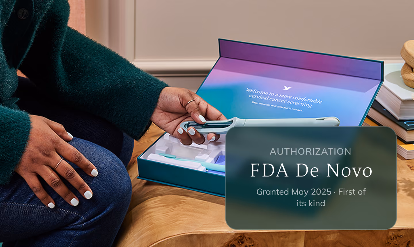 A woman wearing a dark green sweater and jeans sits on a couch, reaching out her hand to lift the Teal Wand self-collection device from its opened Teal kit box. The premium packaging rests on a wooden coffee table alongside decorative books and a small flower vase.