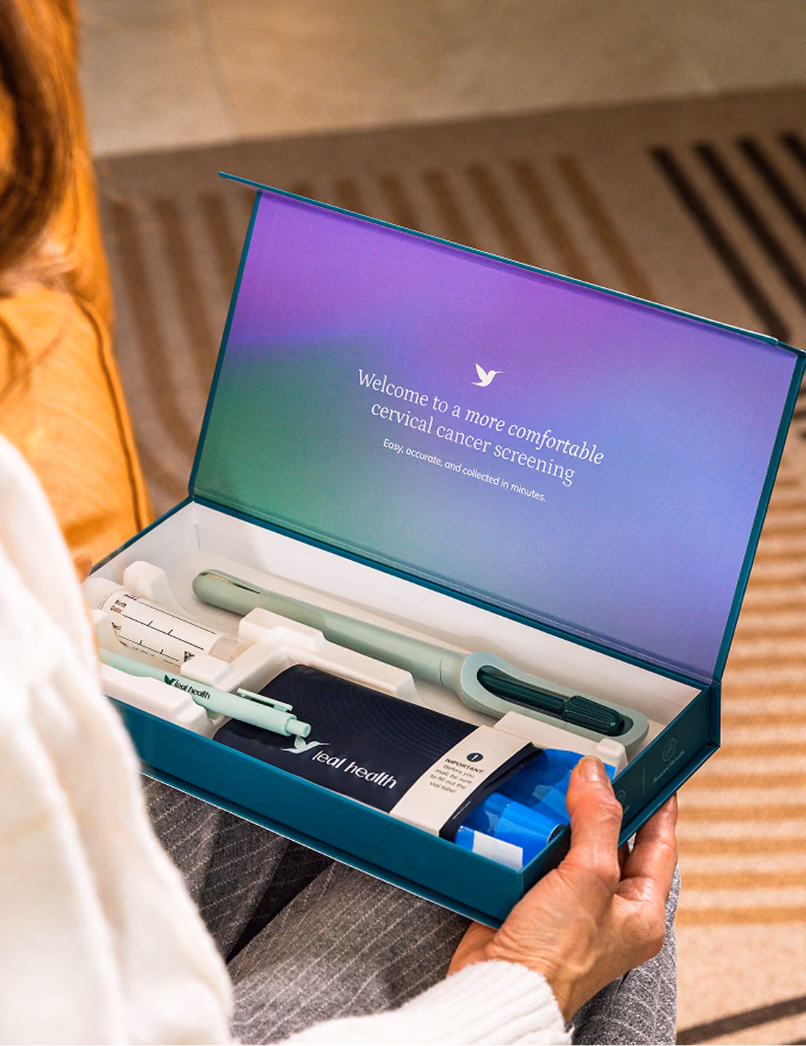 An over-the-shoulder view of a woman unboxing her Teal Health at-home cervical cancer screening kit. The opened box displays the Teal Wand device, a collection vial, a return mailer envelope, and an inside lid reading "Welcome to a more comfortable cervical cancer screening."