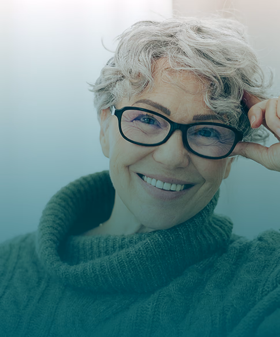 A smiling mature woman with short grey hair and glasses looks directly at the camera while wearing a cozy green cowl-neck sweater, representing healthy and proactive screening practices at every age.
