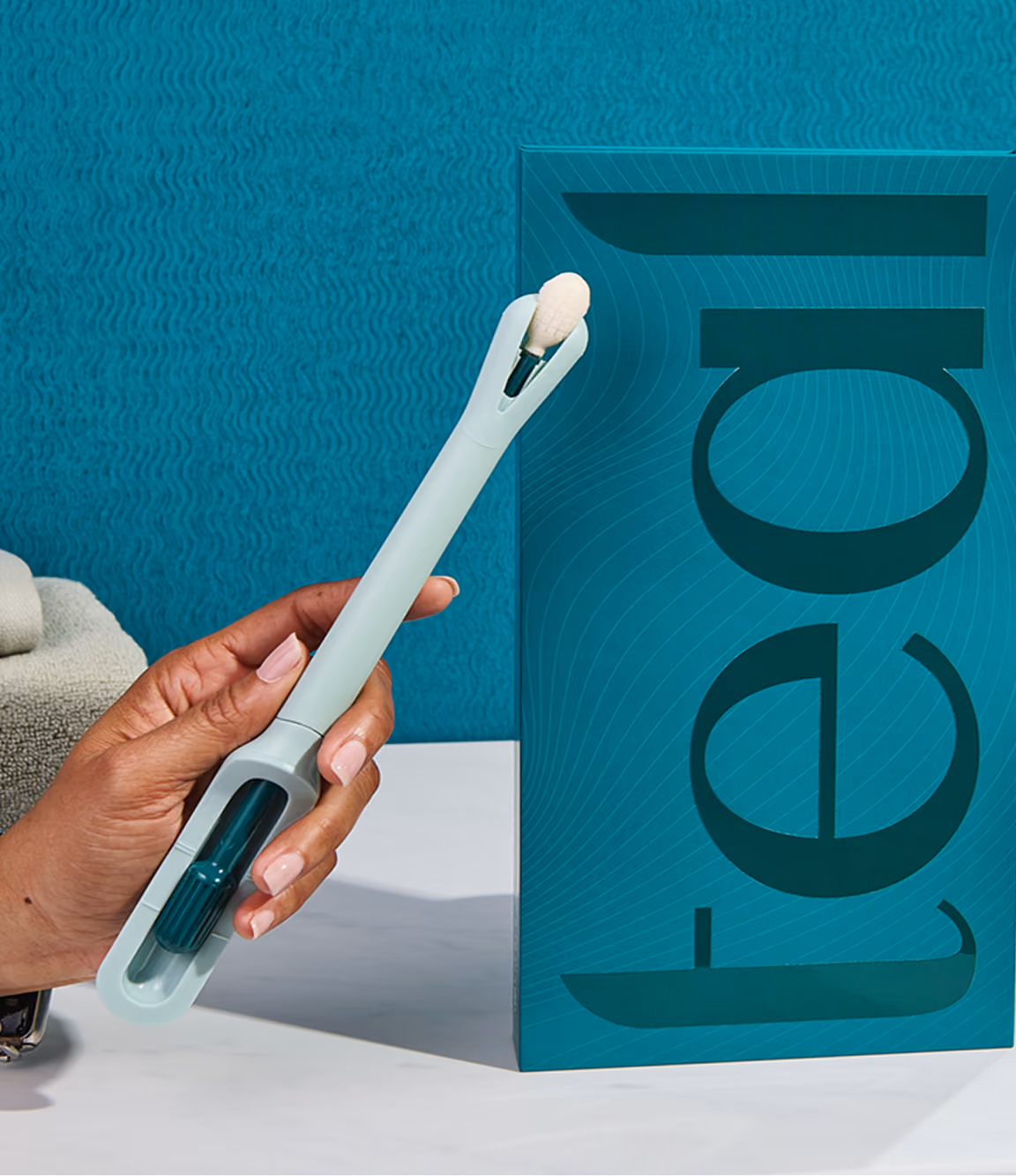 A hand holds up the light-teal colored Teal Wand self-collection device next to its vertical product packaging on a white marble bathroom vanity, showcasing its modern, user-friendly design.
