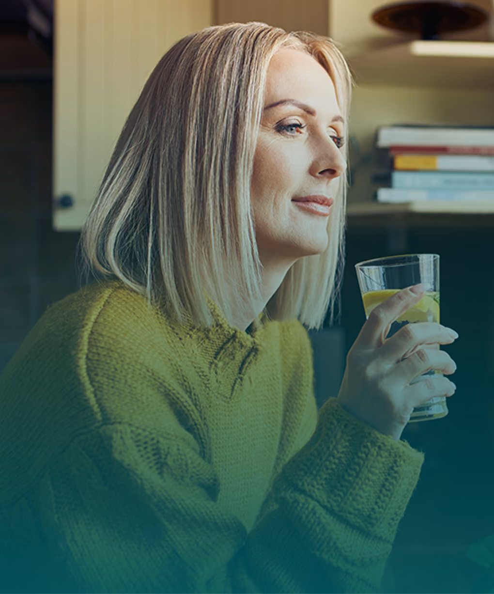A woman looking out a window while holding a glass of water, surrounded by indoor house plants in a warm kitchen setting.