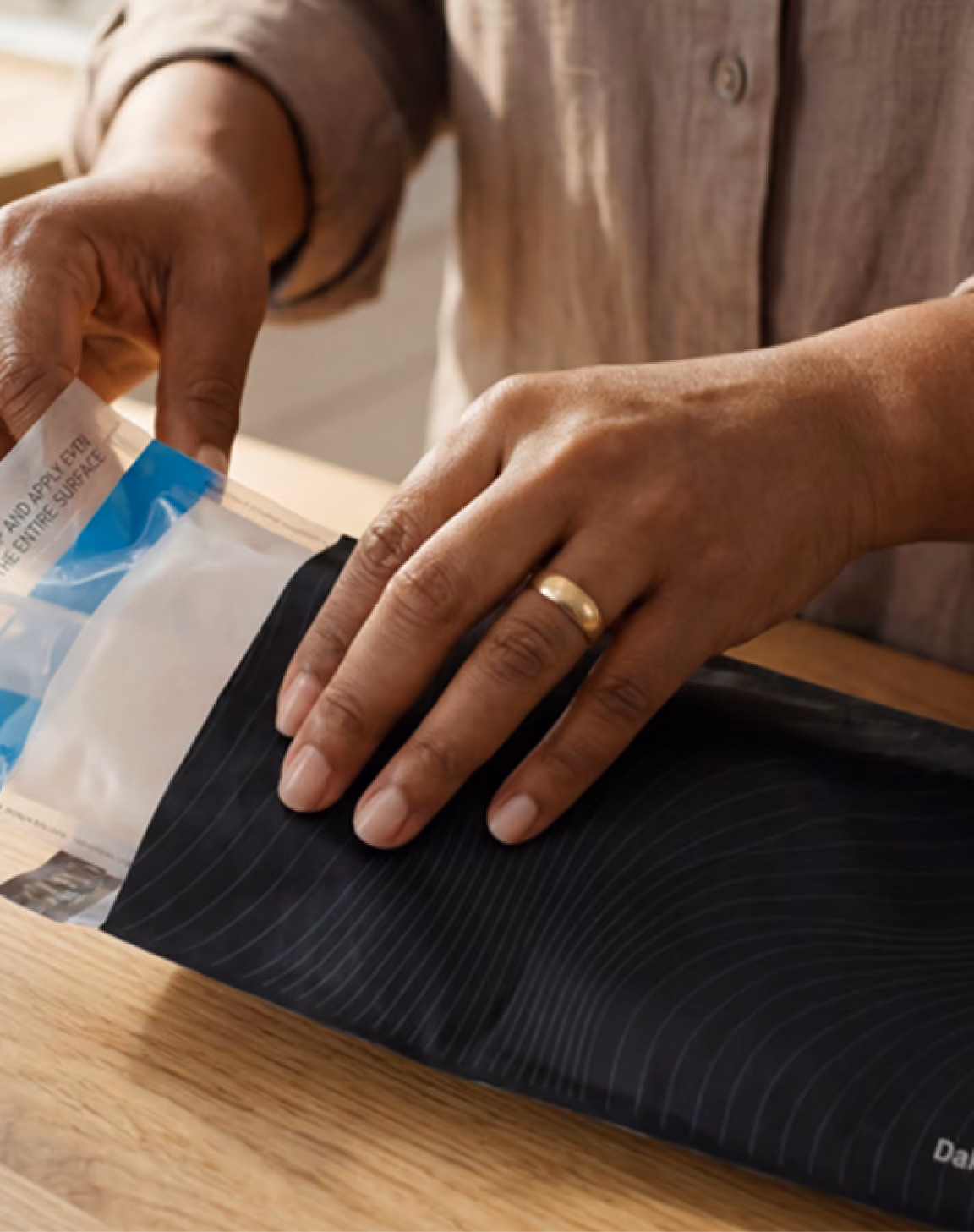Hands with a gold ring pressing a black mailer envelope on a wooden table beside a small labeled instruction pouch.