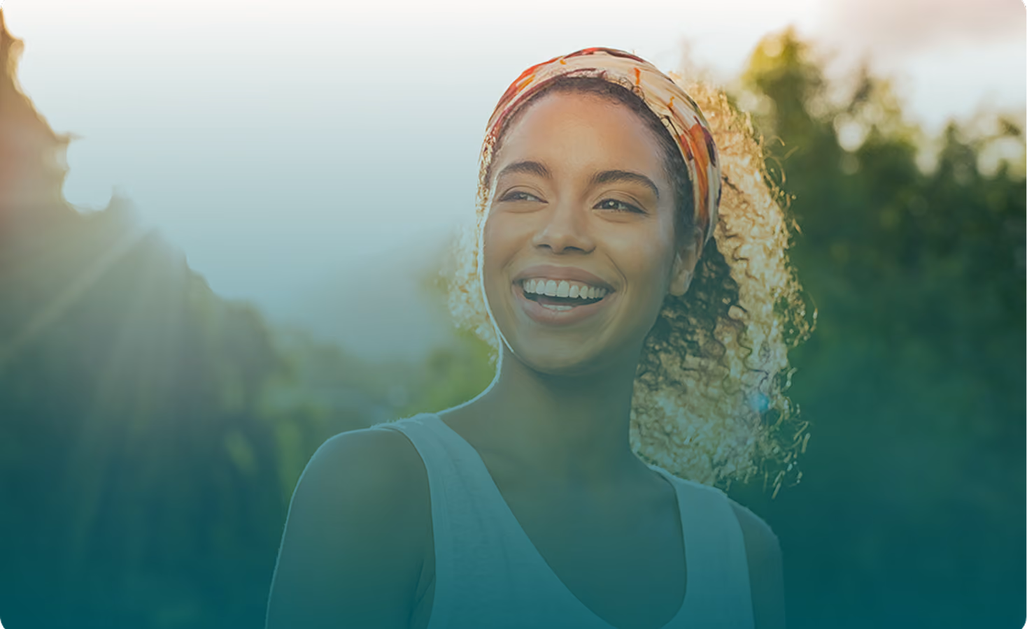 Young woman with a headscarf and tank top smiling outdoors in warm sunlight with greenery behind her.