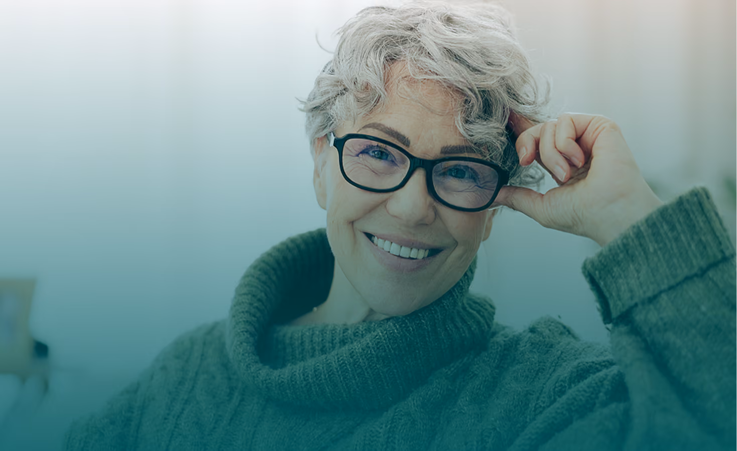 A smiling mature woman with short grey hair and glasses looks directly at the camera while wearing a cozy green cowl-neck sweater, representing healthy and proactive screening practices at every age.