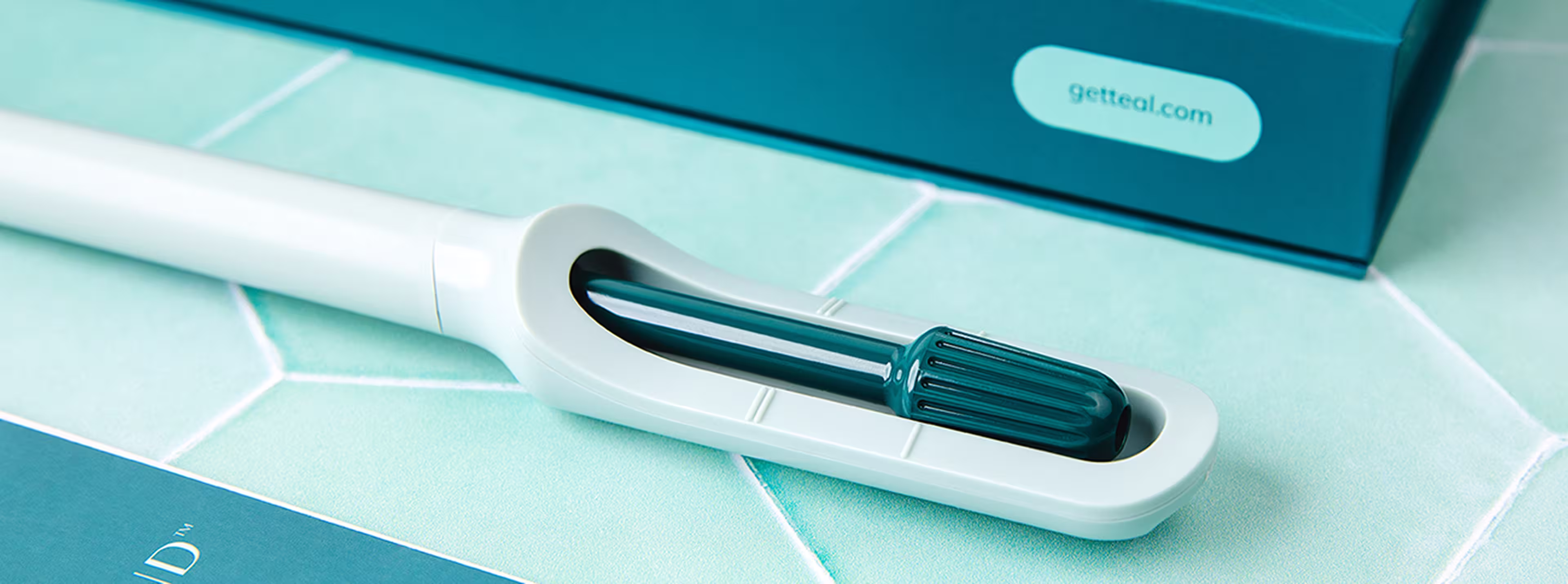 The white and teal Teal Wand device on a green tiled surface beside a teal box labeled getteal.com.